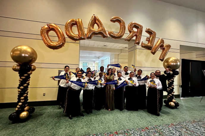 Texas Wesleyan's Mariachi Oro Azul in full getup standing in front of a balloon banner that reads Mercado